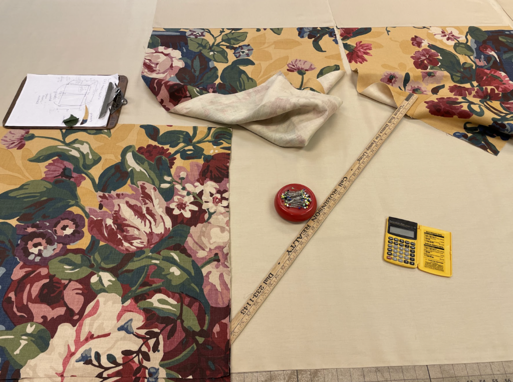 Floral fabric, measuring stick, calculator, pincushion, and clipboard with notes are arranged on a beige work surface.