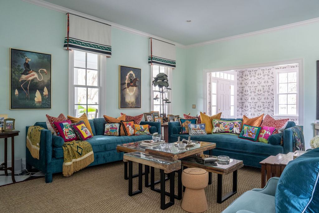 Living room with two blue sofas, colorful pillows, framed art on light blue walls, nested coffee tables, and large windows with white blinds.
