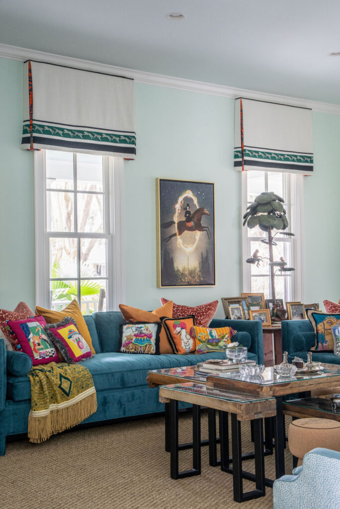 A living room with teal sofas, colorful cartoon pillows, a wooden coffee table, framed photos, and a wall art of an animal riding a broom between two tall windows with patterned valances.