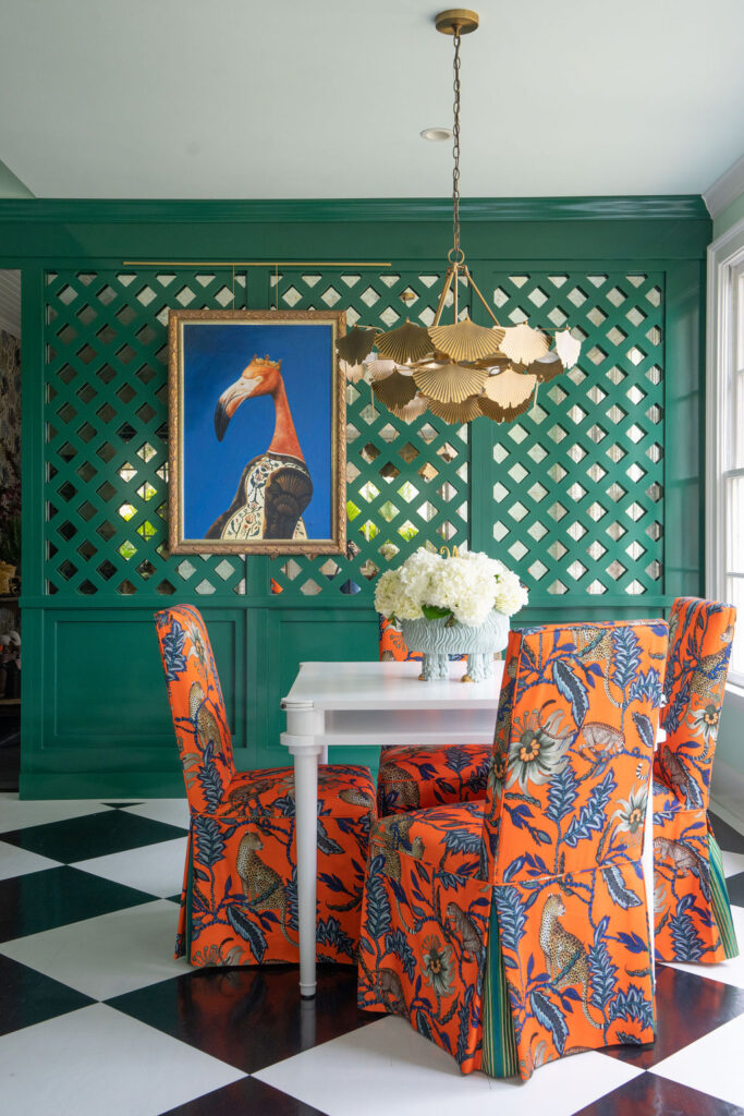 A dining area with orange patterned chairs, a white table, a gold chandelier, green lattice wall, and a framed flamingo artwork; black and white checkered floor.