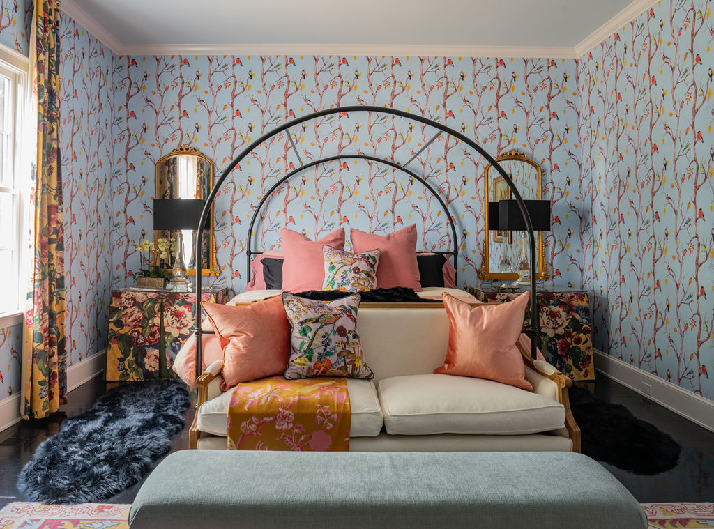 A bedroom with floral wallpaper, an arched black bed frame, pink and floral pillows, mirrored nightstands, black lamps, a white sofa, and a blue upholstered bench.