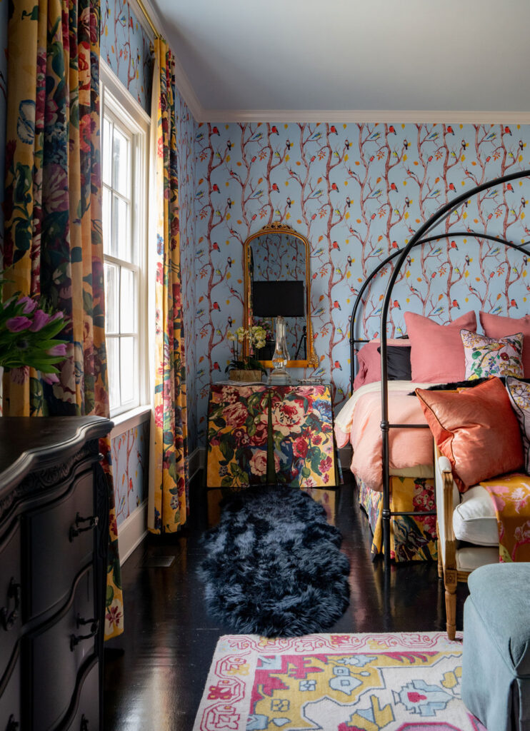 Bedroom with floral wallpaper, colorful bedding, patterned curtains, a black iron bed frame, decorative pillows, a mirror, and a black furry rug on a dark floor.