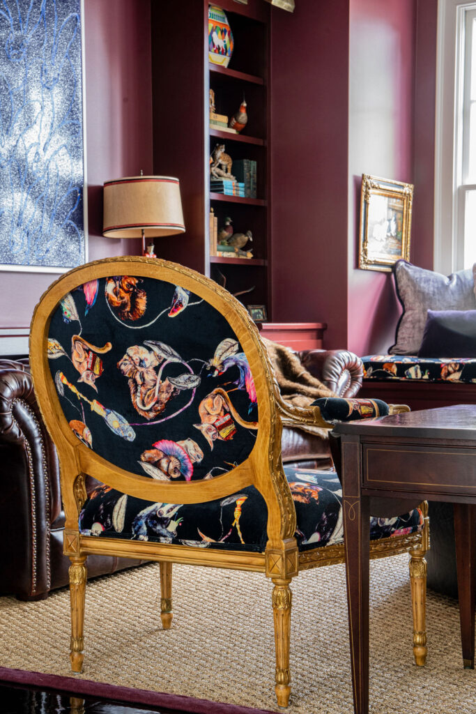 Ornate wooden chair with black upholstery featuring colorful animal designs in a richly decorated room with purple walls and bookshelves.