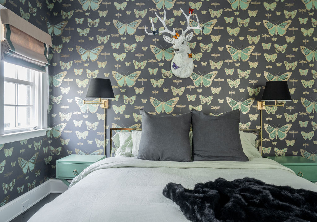 Bedroom with butterfly-patterned wallpaper, a bed with gray pillows and light bedding, black wall lamps, mint green nightstands, and a white deer head wall decoration above the headboard.
